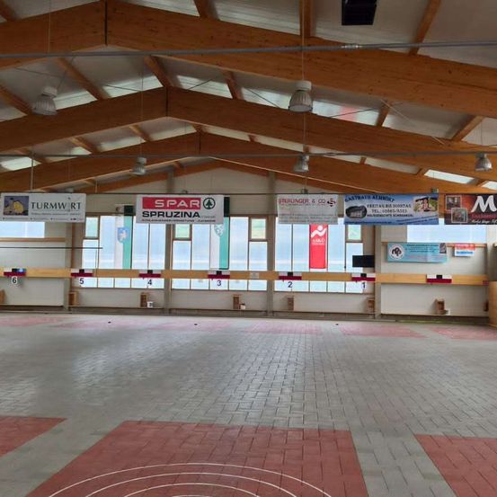 Ein Indoor-Bereich mit Ziegelboden, leerem Raum, hölzerner Dachkonstruktion, hängenden Lampen, Bannern mit Nummern und einem großen Glasfenster.