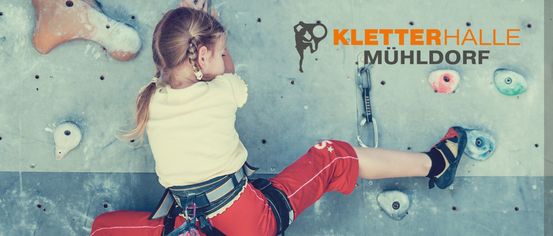 Eine Person klettert an einer Kletterwand mit dem Text Kletterhalle Mühldorf. Sie trägt einen Klettergurt und Schuhe.