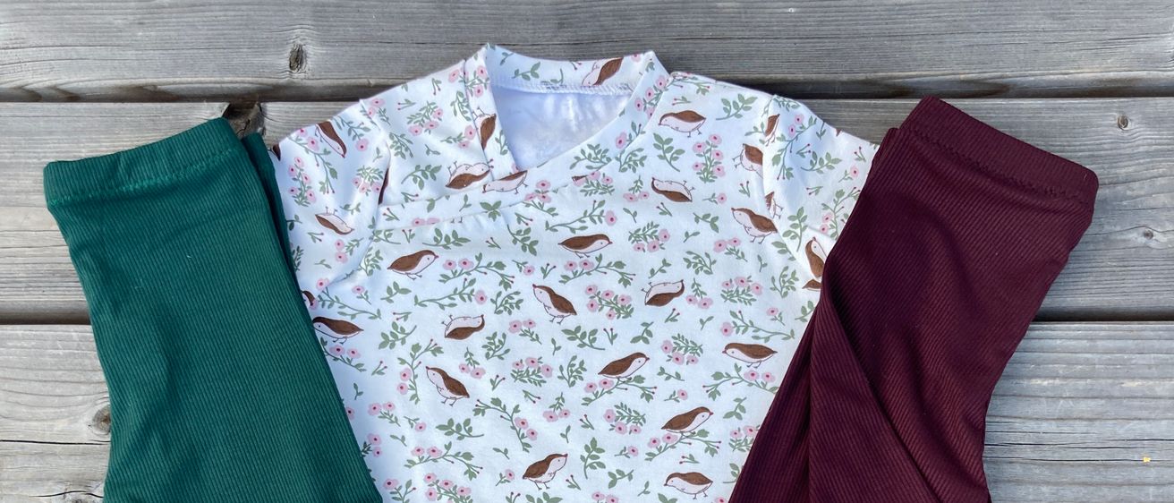 Ein weißes Shirt mit Vogel-Print und zwei gerippte Hosen, eine grün und eine bordeauxrot, liegen auf einer Holzoberfläche.