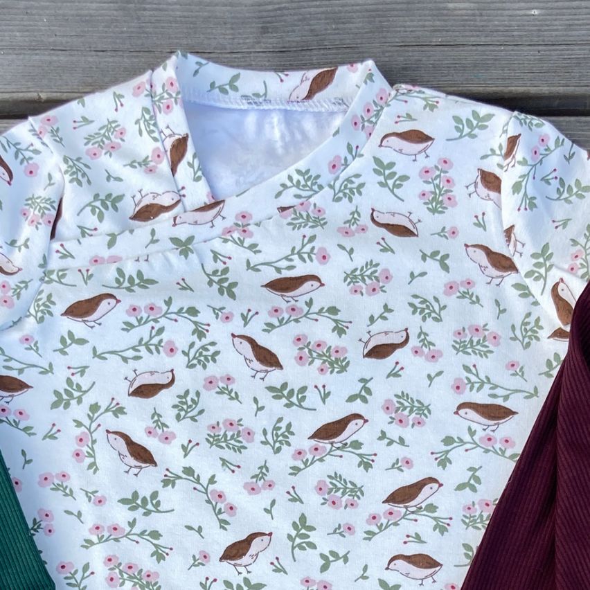 Ein weißes Shirt mit Vogel-Print und zwei gerippte Hosen, eine grün und eine bordeauxrot, liegen auf einer Holzoberfläche.