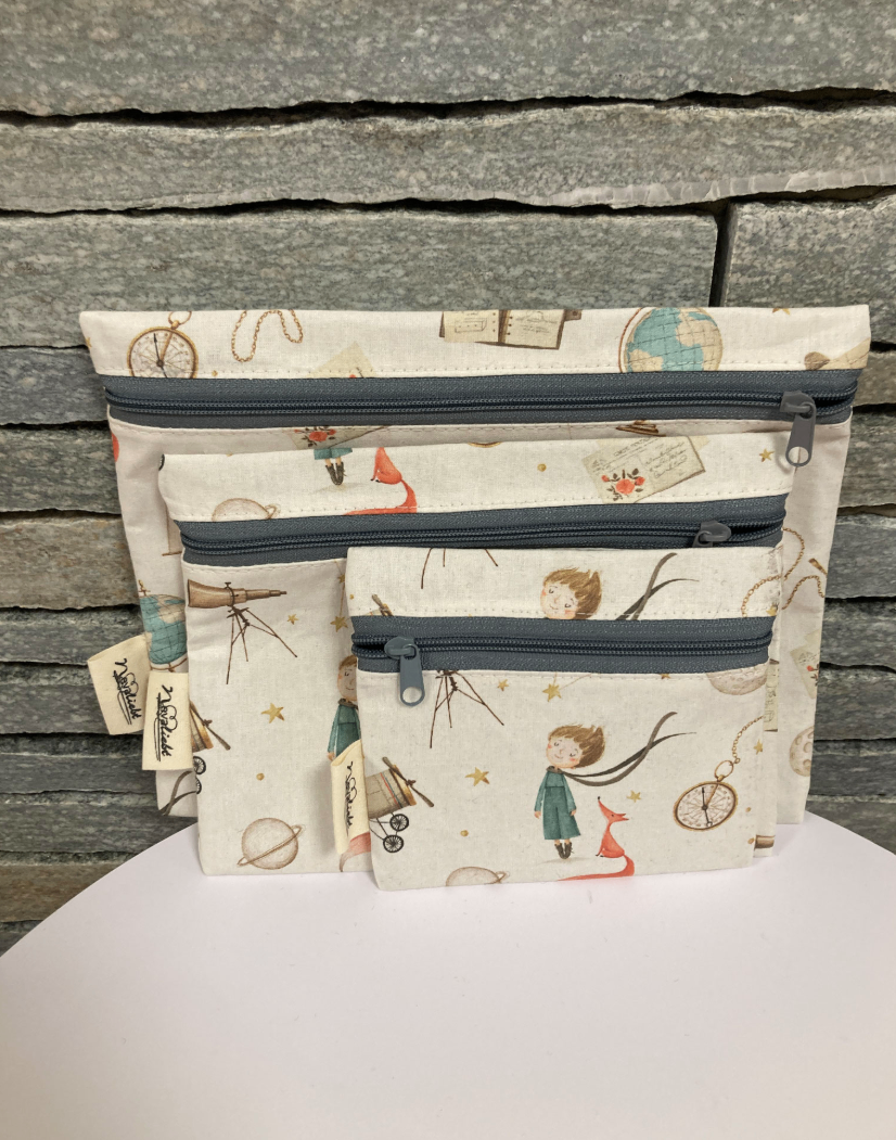 Drei gezippte Taschen mit Weltraumthema liegen auf einer weißen Oberfläche. Die größte Tasche hat eine Erdkugel und andere Gegenstände, die mittlere zeigt ein Teleskop und die kleinste zeigt ein Mädchen mit einem Fuchs.