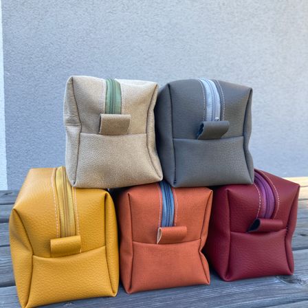 Fünf kleine Ledertaschen in verschiedenen Farben auf einer Holzoberfläche gestapelt. Die Taschen sind hellbraun, grau, orange, gelb und rot. Jede Tasche hat einen Reißverschluss und einen kleinen Klappverschluss.