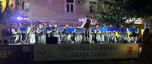 Ein Blechbläserensemble spielt nachts auf der Bühne vor Gebäuden mit vielen Fenstern.