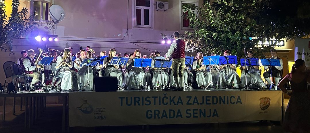 Ein Blechbläserensemble spielt nachts auf der Bühne vor Gebäuden mit vielen Fenstern.