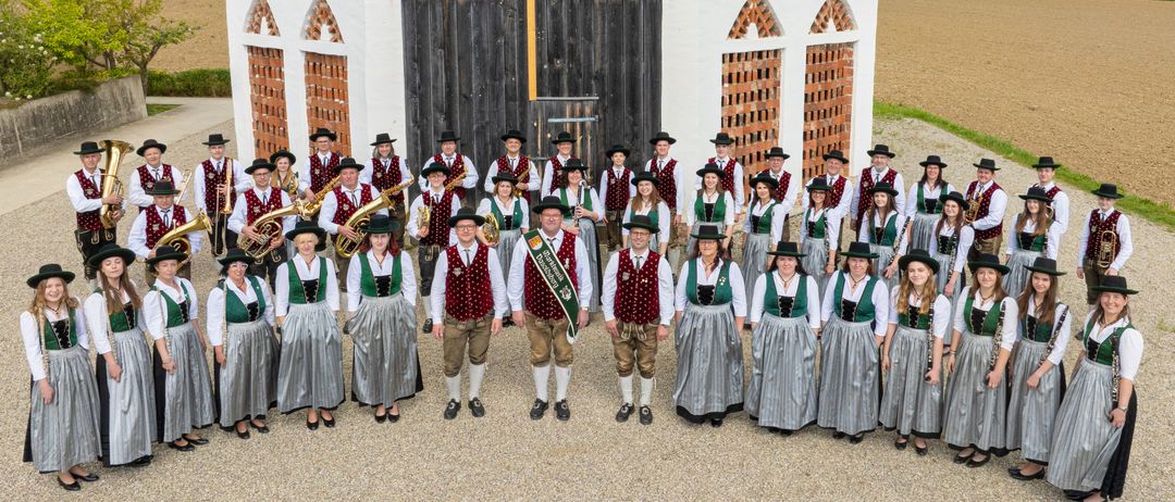 Eine Gruppe von Musikern in traditioneller deutscher Kleidung steht vor einem historischen Gebäude mit Ziegeldetails. Das Gebäude befindet sich in einer ländlichen Umgebung mit einem großen Feld und Bergen in der Ferne.