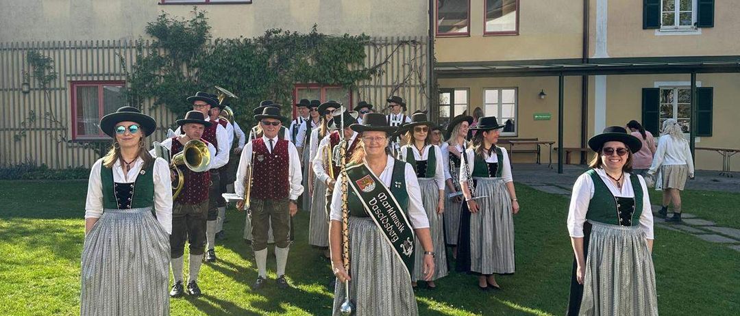 Ein Blasorchester in traditioneller deutscher Kleidung steht auf einem Rasen vor einem historischen Gebäude mit roten Rahmen. Sie halten Musikinstrumente in der Hand.