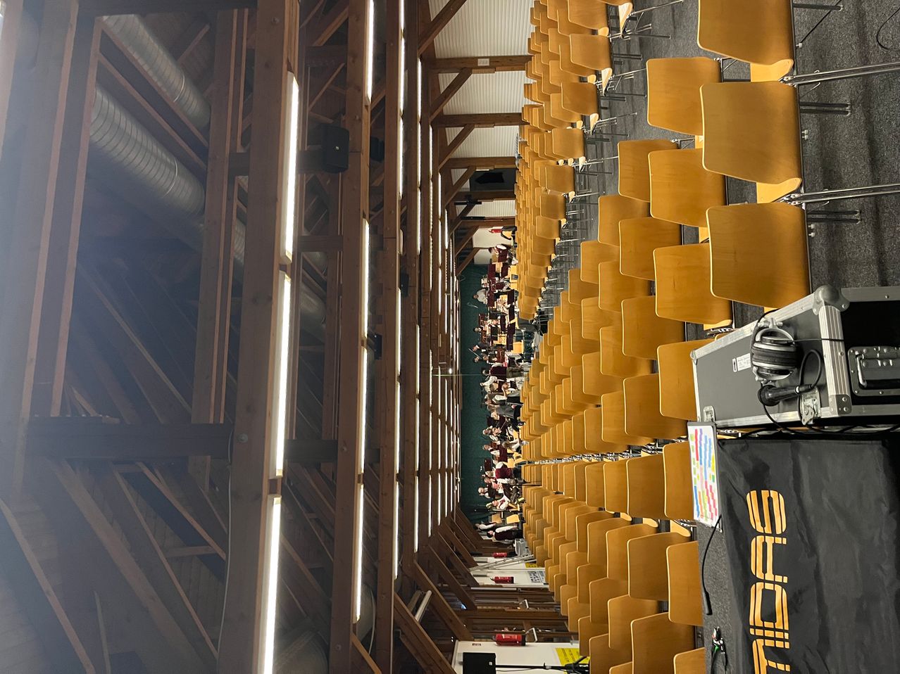 Ein großer Saal mit Holzstühlen, Holzbalken und einer Bühne, auf der Menschen in Reihen sitzen.