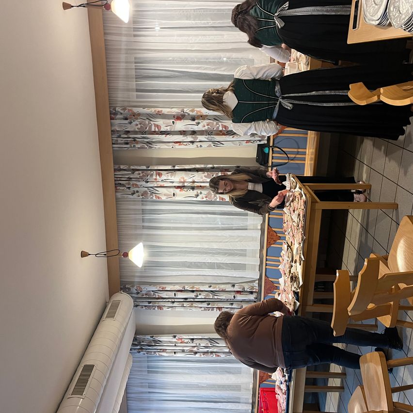 Drei Frauen in grünen und schwarzen Kleidern stehen über einem Esstisch. Eine schaut sich das Essen an, eine andere hält eine Tasche. Ein Mann in einem braunen Hemd lehnt sich über den Tisch. Der Raum hat Holzstühle, einen weißen Vorhang und eine weiße Klimaanlage.