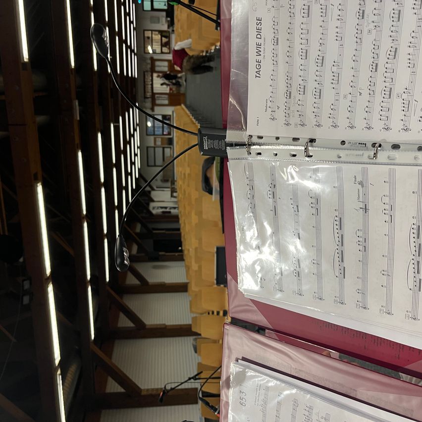 Ein Notenblatt mit dem Titel 'Tage wie diese' befindet sich in einem transparenten Ordner in einem roten Ordner. Der Raum hat einen Holzboden und eine Decke mit Lichtern.