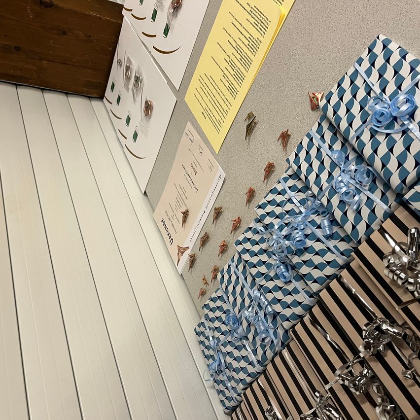 Eine Reihe von ordentlich verpackten Geschenken, einige mit blauen Schleifen, ist vor einem grauen Hintergrund ausgestellt. Papiere mit Text und Bildern sind an der Wand angeheftet.