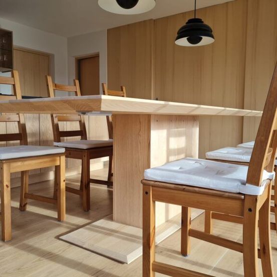 Ein Esszimmer mit hölzernen Stühlen um einen großen Holztisch, hölzerne Wände und ein weißer Boden. Zwei Lampen hängen von der Decke.