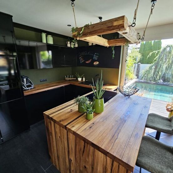 Eine moderne Küche mit einer großen hölzernen Insel, grünen Pflanzen und Blick auf einen Pool draußen. Die Küche hat schwarze Schränke und eine Kreidetafel mit Speisekarte.