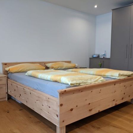 Ein Schlafzimmer mit einem Holzbett, blauen Laken und gelben Kissen. Ein hölzener Nachttisch ist links. Zwei graue Schränke sind rechts.