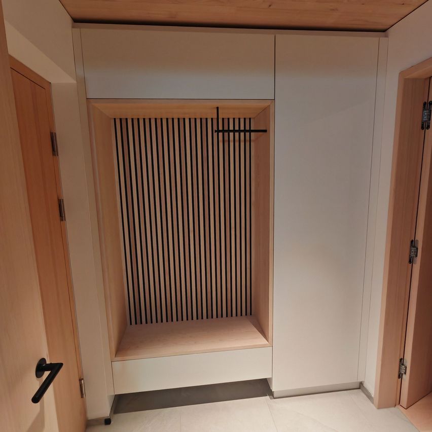 Innenraum eines Raumes mit weißen Wänden und einem Holzboden. Ein Holzschrank mit weißer Tür, einer Bank und einem schwarzen Metallkreuz steht in der Mitte. Zwei Holztüren befinden sich auf beiden Seiten.