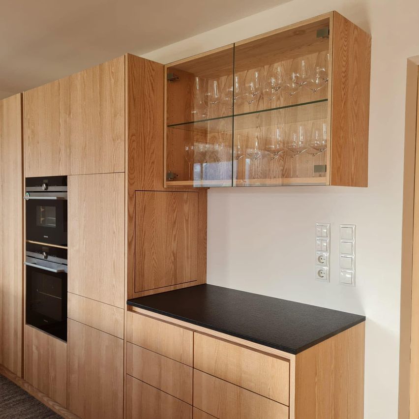 Eine moderne Küche mit Holzschränken und einer schwarzen Arbeitsplatte. Die Schränke haben Glastüren und Regale für Weingläser. Die Wand hat Schalter und eine Steckdose.