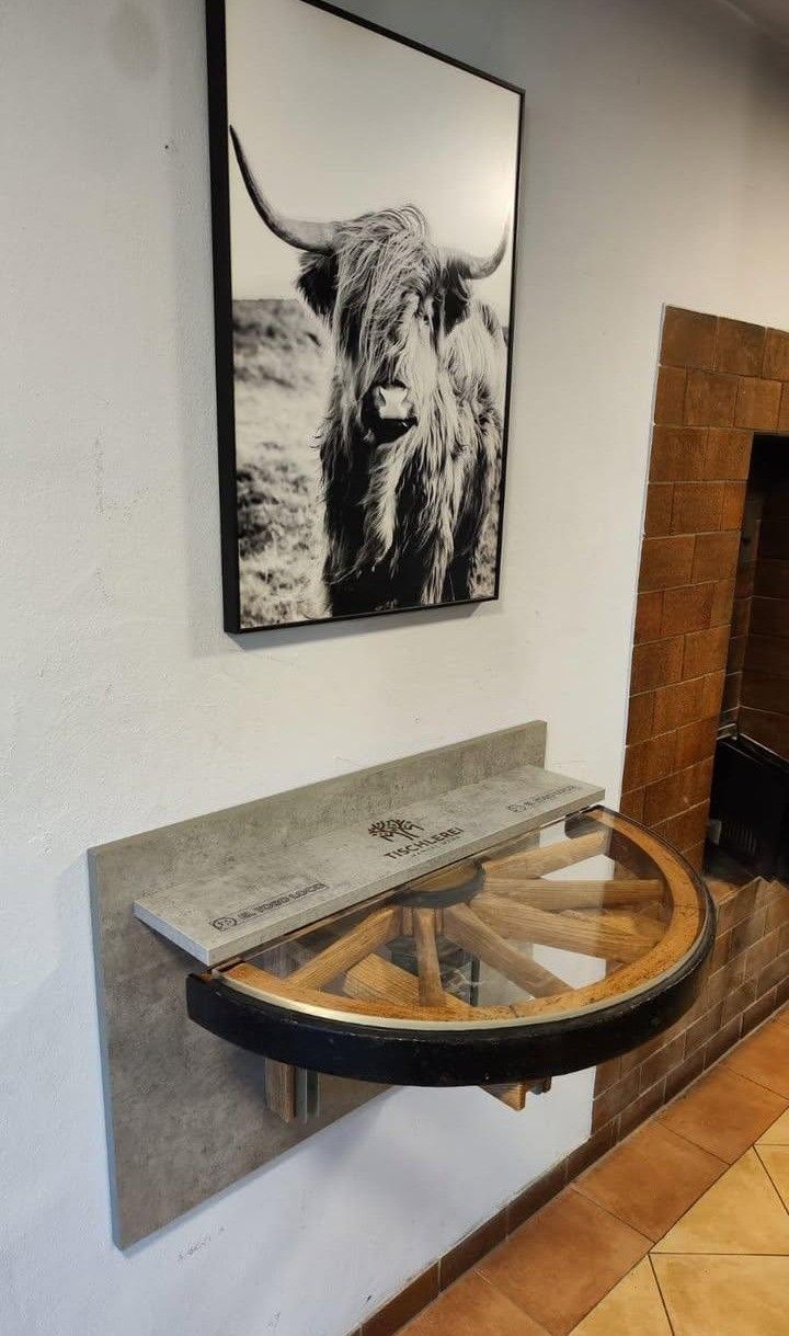 Eine Wand mit einem gerahmten Foto eines Highland-Rinds. Daneben ein runder Tisch mit Glasplatte und hölzerne Räder. Der Tisch hat eine graue Betonplatte obenauf.
