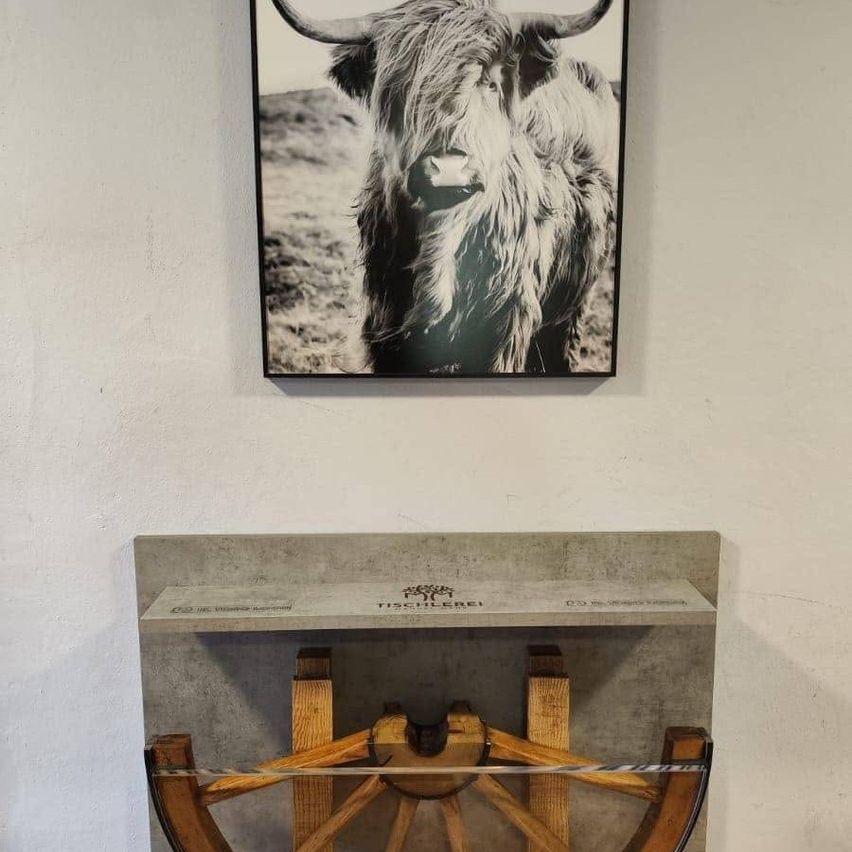 Ein an der Wand befestigtes Foto eines Hochlandrindes über einer Betonbank mit einem Holzrad, mit der Marke Tischlerrei auf der Oberseite.