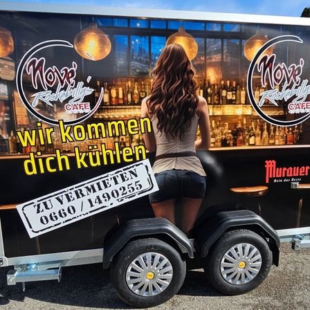 Eine Frau mit langen Haaren und einem Gürtel steht auf einem Anhänger mit einer Bartheke im Hintergrund. Der Anhänger hat zwei große Räder und ein Nummernschild mit einer Telefonnummer und 'ZU VERMIETEN' darauf. Die Bartheken haben Regale gefüllt mit Likörflaschen. Der Name der Bar ist in Deutsch als 'Nove's Rockabilly Cafe' geschrieben.