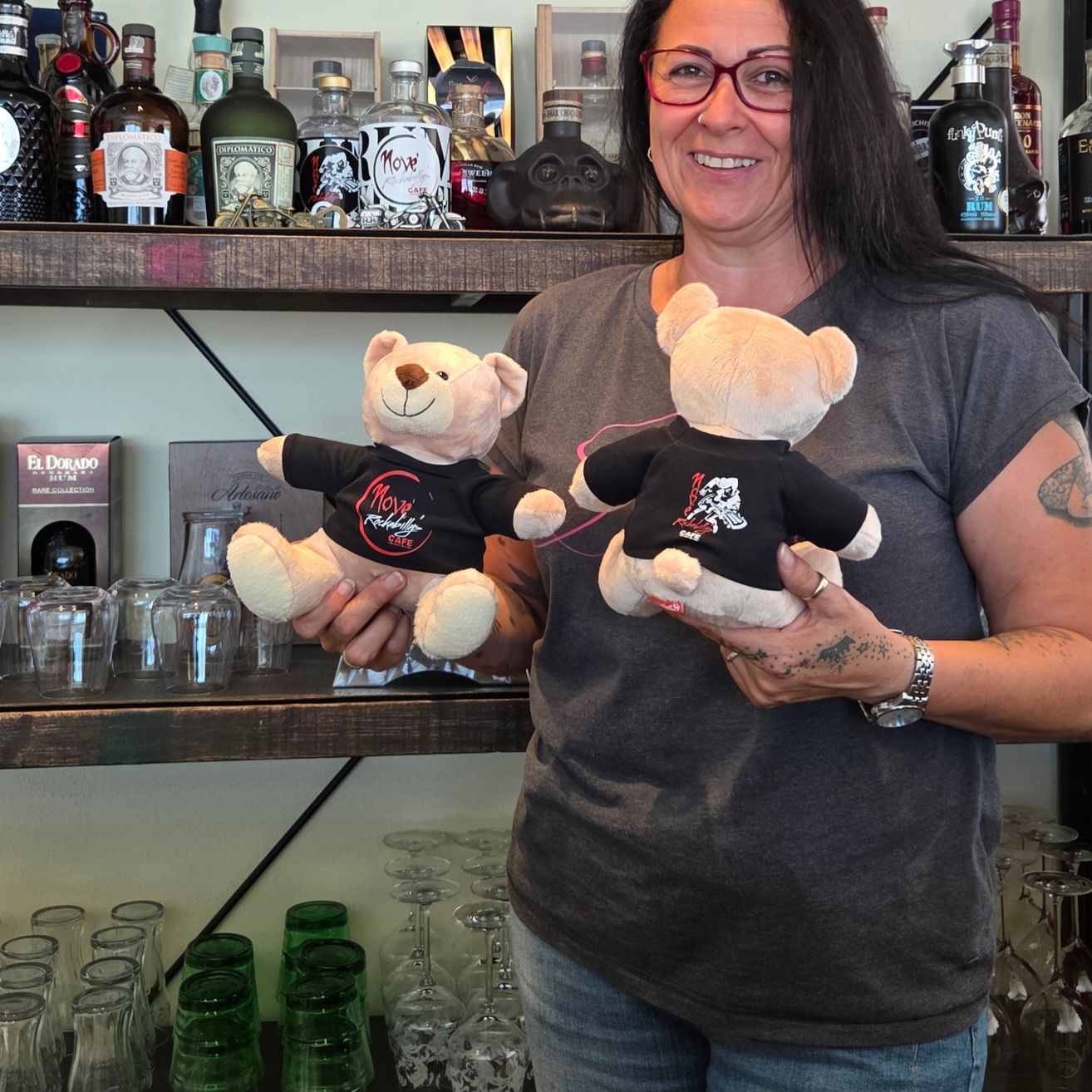 Eine Frau steht vor einem Regal mit verschiedenen Likörflaschen, lächelt und hält zwei Teddybären, die schwarze T-Shirts mit dem Wort Move tragen.