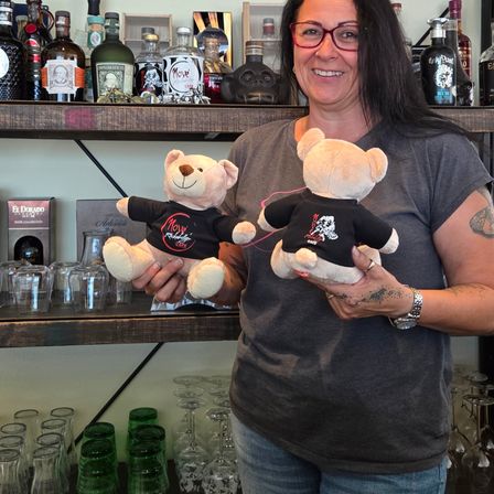 Eine Frau steht vor einem Regal mit verschiedenen Likörflaschen, lächelt und hält zwei Teddybären, die schwarze T-Shirts mit dem Wort Move tragen.