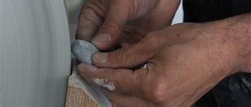 Ein Mann formt ein Tonobjekt auf einer Drehscheibe mit einem Stein. Er trägt ein schwarzes Hemd und hat einen Ring am Finger.