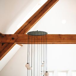 Eine moderne Pendelleuchte mit Glasbirnen hängt von Holzbalken an einer hohen Decke. Der Raum hat weiße Wände und Decke, mit einfallendem Sonnenlicht.