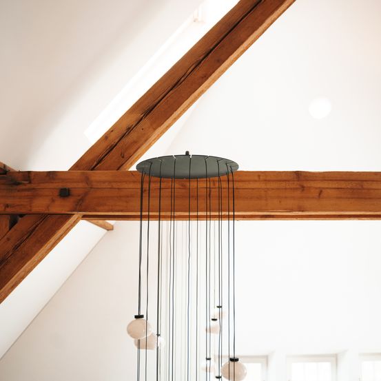 Eine moderne Pendelleuchte mit Glasbirnen hängt von Holzbalken an einer hohen Decke. Der Raum hat weiße Wände und Decke, mit einfallendem Sonnenlicht.