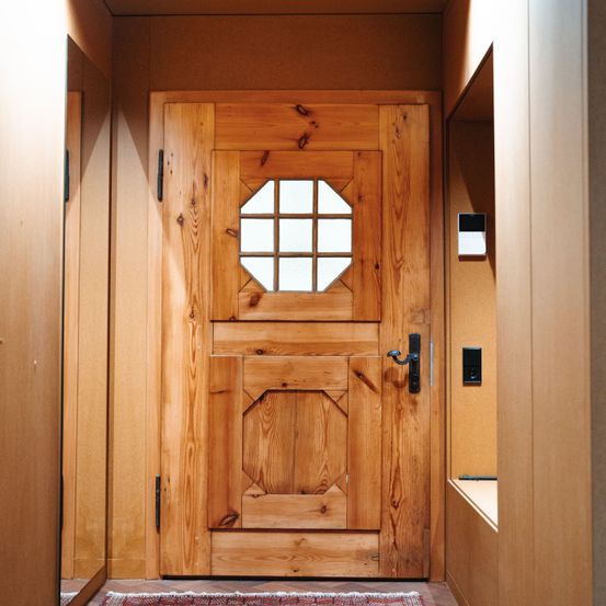 Eine Holztür mit einem sechseckigen Fenster ist an einer Holzwand montiert. Die Tür ist leicht geöffnet, und ein Teppich liegt auf dem Boden.