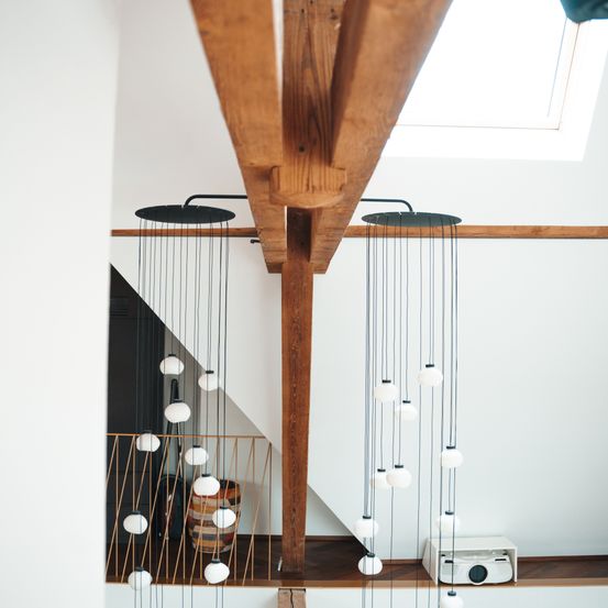 Ein Innenansicht einer Holzbalkendecke mit hängenden Lampen und einem Beamer. Der Bereich ist gut beleuchtet und die Lampen sind leuchtend. Ein Holzgeländer ist sichtbar.