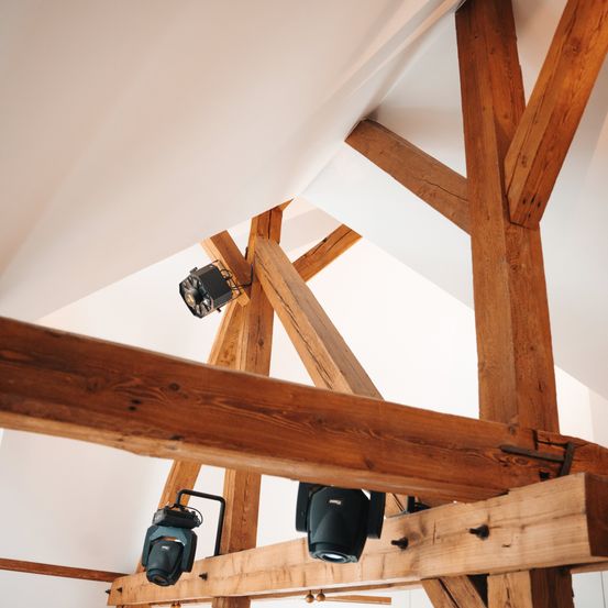 Eine Holzkonstruktion mit Balken, die eine Decke stützen, mit zwei schwarzen Strahlern befestigt. Die Holzbalken sind sichtbar und die Decke ist weiß.