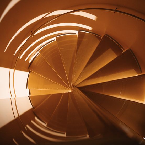 Eine Nahaufnahme einer Wendeltreppe mit goldbraunen Stufen, die Licht von oben reflektiert.