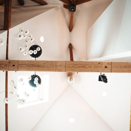 Eine Holzbalkendecke mit einer weißen Wand, an der mehrere kreisförmige Lichter hängen. Eine schwarze kreisförmige Leuchte ist an der Decke montiert. Ein Fenster befindet sich unter der Decke.