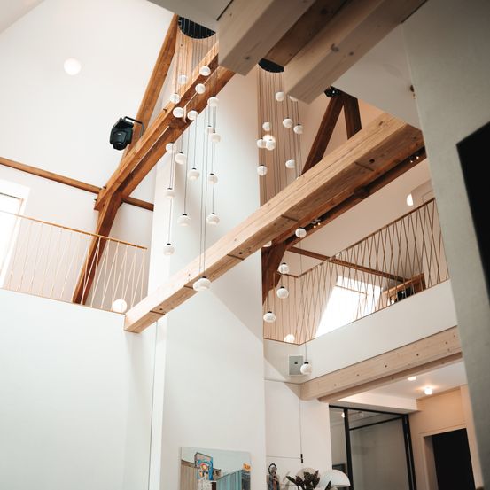Ein Innenansicht eines Hauses mit Holzbalken und Lichterketten, die von der Decke hängen. Zwei Balkone mit Geländern sind sichtbar.