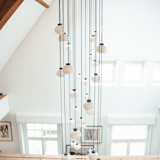 Ein modernes, minimalistisches Interieur mit einer Pendelleuchte aus weißen Glaskugeln, die an der Decke hängt. Der Raum hat weiße Wände, Holzbalken und große Fenster. Gerahmte Bilder hängen an der Wand unter den Fenstern.