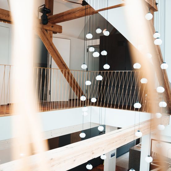 Ein modernes architektonisches Interieur mit Holzbalken und hängenden Leuchten. Der Raum verfügt über eine Glaswand, einen Holzboden und einen Balkon mit einer Brüstung.