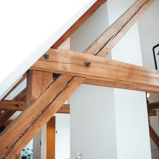 Ein Nahaufnahme-Blick auf Holzbalken im Innenraum eines Hauses. Die Balken sind mit Metallbefestigungen verbunden. Im Hintergrund befindet sich eine weiße Wand.