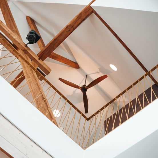 Eine Holzdecke mit Balken und einem Deckenventilator. Zwei Lichter sind an der Decke. Eine Kamera ist links und ein Holzgeländer ist rechts.