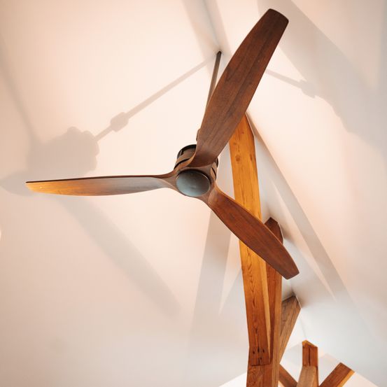 Ein Holzventilator ist an einer weißen Decke montiert. Der Ventilator hat eine hölzerne Klinge und eine schwarze Mitte. Er wirft einen Schatten an die Wand.