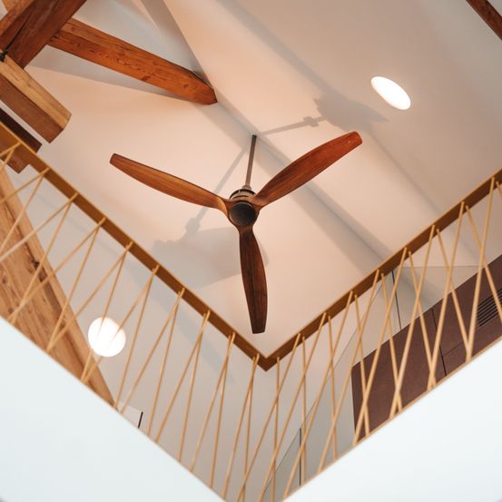 Ein Holzdeckenventilator mit Flügeln hängt von einer weißen Decke, über einem hölzernen Geländer mit Glasscheibe. Eine runde Leuchte ist an der Decke angebracht.