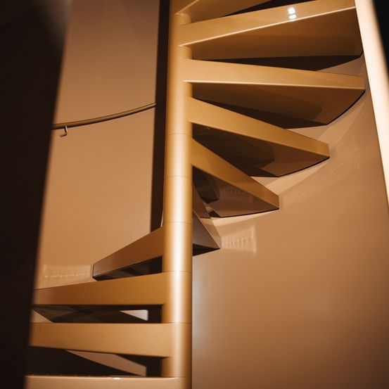 Eine Nahaufnahme einer goldfarbenen Wendeltreppe mit einem Metallgeländer an einer beigefarbenen Wand.