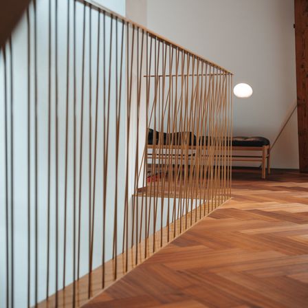 Ein modernes Interieur mit weißer Wand, Holzboden und Messinggeländern. Eine kleine weiße Lampe befindet sich an der Decke. Eine Holzbank mit Kissen steht an der Wand.
