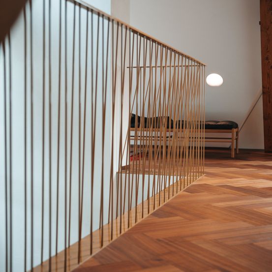 Ein modernes Interieur mit weißer Wand, Holzboden und Messinggeländern. Eine kleine weiße Lampe befindet sich an der Decke. Eine Holzbank mit Kissen steht an der Wand.