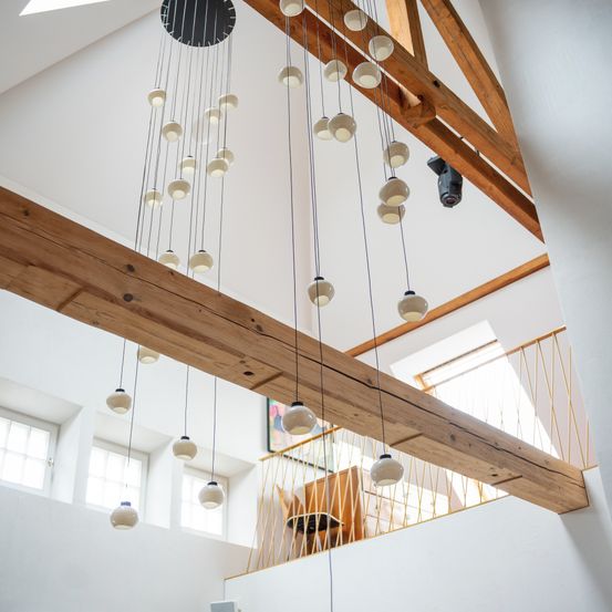 Ein moderner Raum mit Holzbalken, weißen Wänden und einer Hängeleuchte. Eine Treppe mit Geländer führt zu einem Balkon mit einem Schreibtisch.