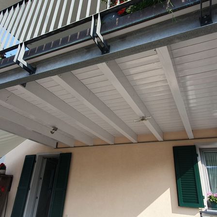 Ein Balkon mit einem weißen Geländer, einer Metallstütze und einer weißen Decke mit einem Ventilator. Unten ein Gebäude mit einer Tür, einem Fenster und einem Topfpflanzen.