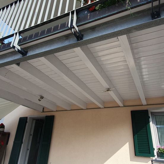 Ein Balkon mit einem weißen Geländer, einer Metallstütze und einer weißen Decke mit einem Ventilator. Unten ein Gebäude mit einer Tür, einem Fenster und einem Topfpflanzen.