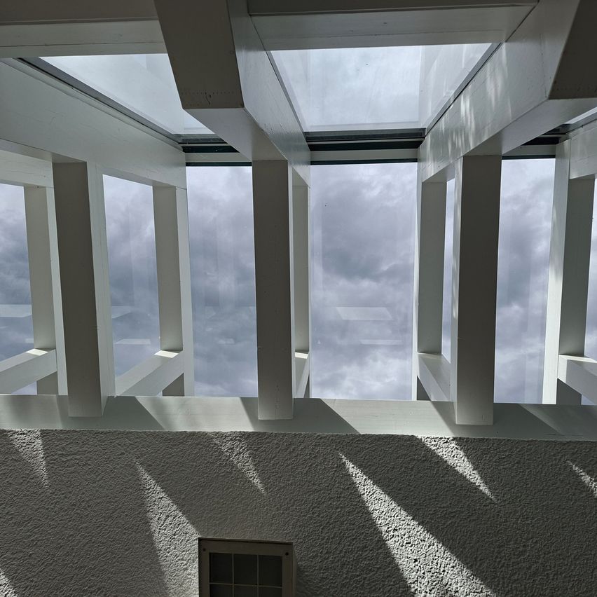 Ein Blick von unten auf eine moderne Glasdachstruktur mit weißen Balken und einem durch das Glas sichtbaren bewölkten Himmel.
