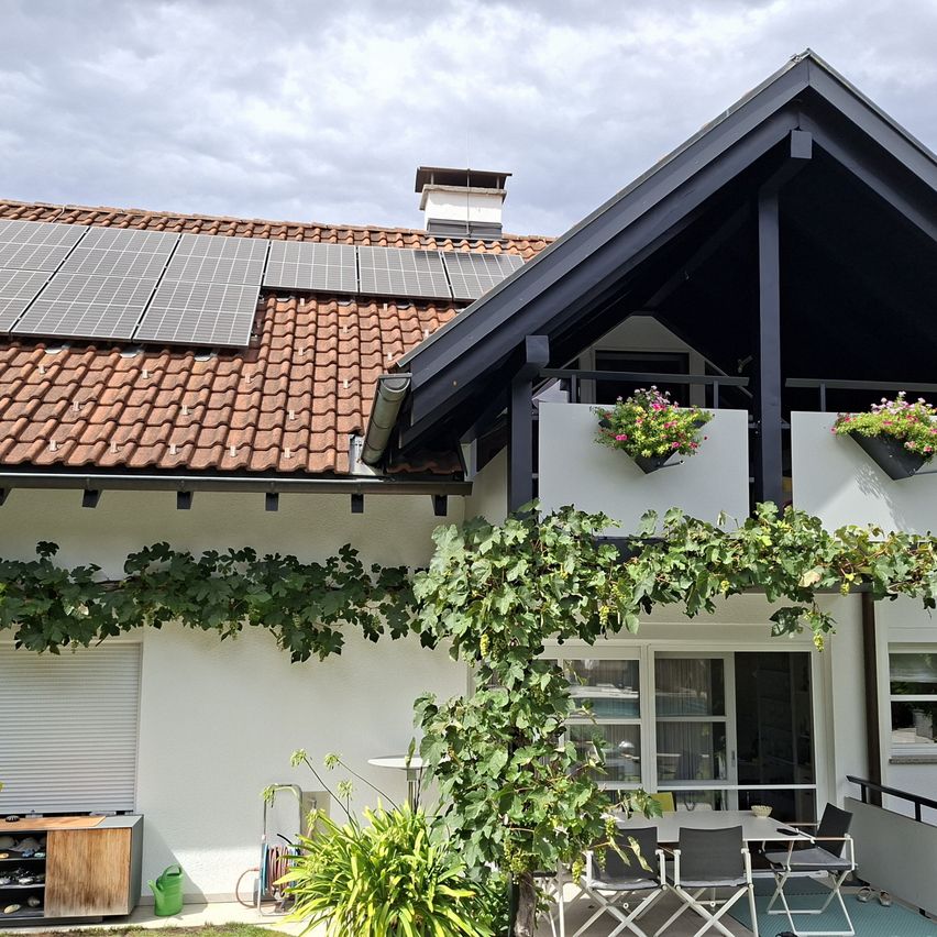 Ein modernes Haus mit einem roten Dach, Solarpanelen und einem Balkon, der mit Topfpflanzen geschmückt ist. Der Vorgarten verfügt über einen Tisch und Stühle unter einer Rebe.