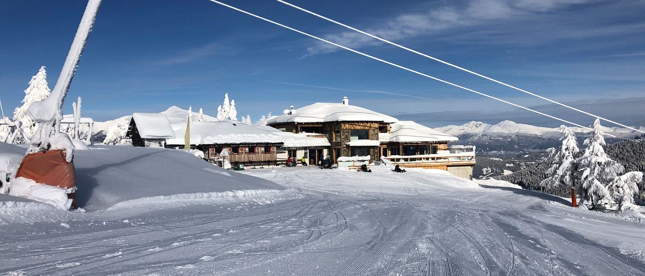 Ein Skigebiet mit mehreren schneebedeckten Gebäuden und Spuren im Schnee. Im Vordergrund ist ein Seilbahnseil zu sehen.