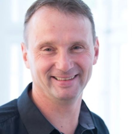 Ein Mann in einem schwarzen Hemd lächelt in die Kamera. Er hat kurze, ordentlich gekämmte Haare und befindet sich in einem gut beleuchteten Raum.