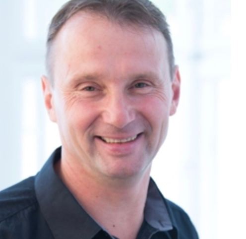 Ein Mann in einem schwarzen Hemd lächelt in die Kamera. Er hat kurze, ordentlich gekämmte Haare und befindet sich in einem gut beleuchteten Raum.